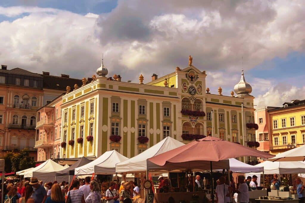 Ein lebhafter Markt im Freien mit vielen Menschen und weißen Zelten vor einem verzierten historischen Gebäude mit Uhr, Kuppeln und dekorativen Details unter einem teilweise bewölkten Himmel.