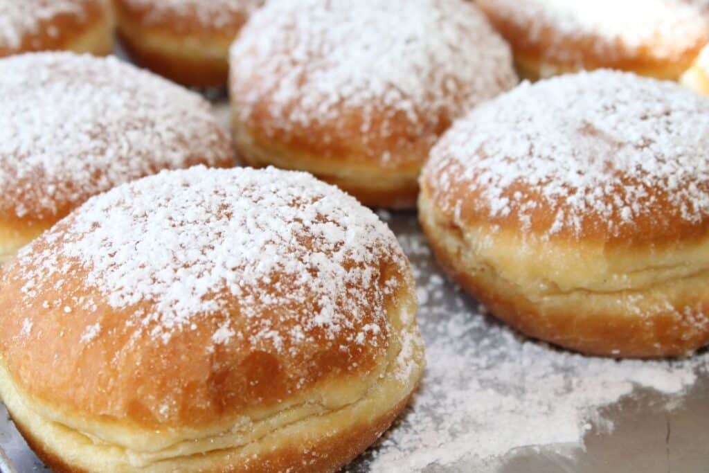 Nahaufnahme mehrerer runder, goldbrauner Donuts, die großzügig mit Puderzucker bestäubt und auf einem Metalltablett angeordnet sind. Die Donuts wirken fluffig und frisch gebacken.