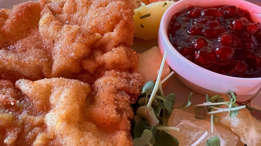 Nahaufnahme eines Tellers mit panierten Schnitzeln, Salzkartoffeln mit Kräutern, einer kleinen Schüssel mit Preiselbeersauce und einer Garnitur aus frischem Grünzeug.