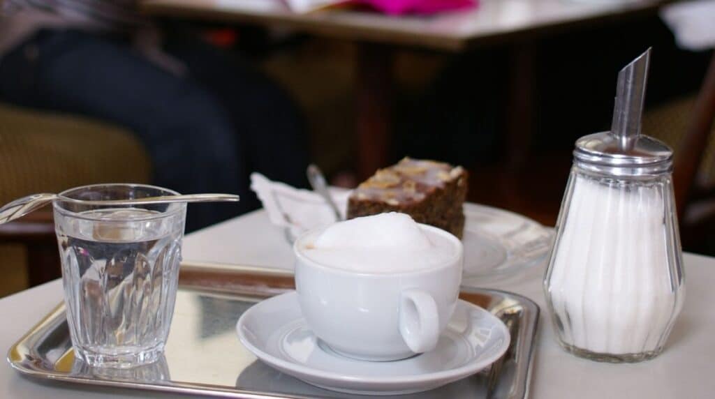 Eine Tasse Cappuccino mit Schaum, ein Glas Wasser und ein Zuckerspender sind auf einem Metalltablett angeordnet. Im Hintergrund sind ein Teller mit einem Stück Kuchen und einige unscharfe Figuren zu sehen, die an einem Tisch sitzen.