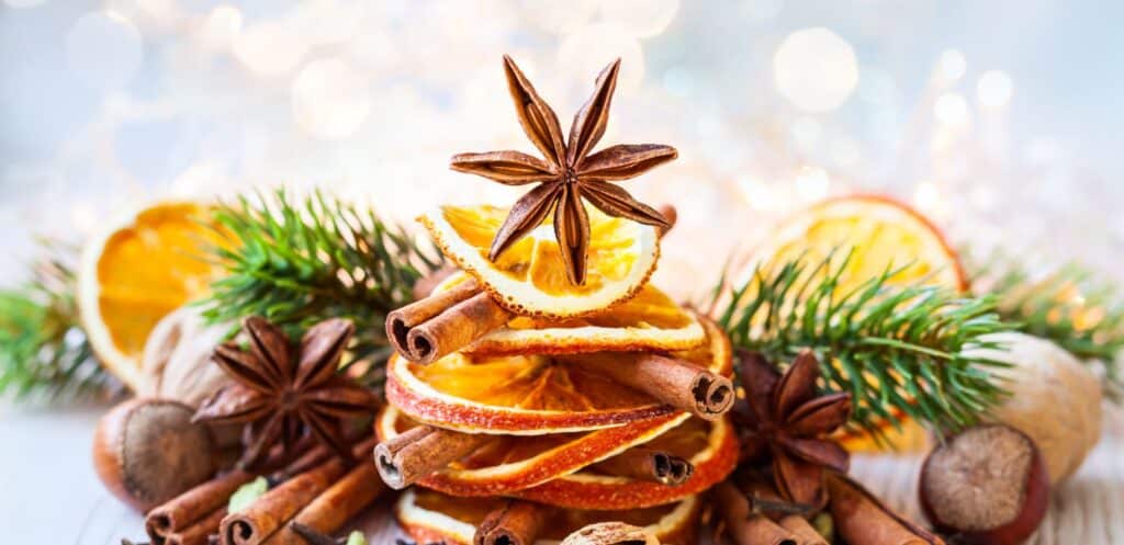 Ein festliches Arrangement aus getrockneten Orangenscheiben, gestapelt mit Zimtstangen, gekrönt mit einem Sternanis, umgeben von Tannenzweigen, Nüssen und Weihnachtsgewürzen auf einem hellen Hintergrund.