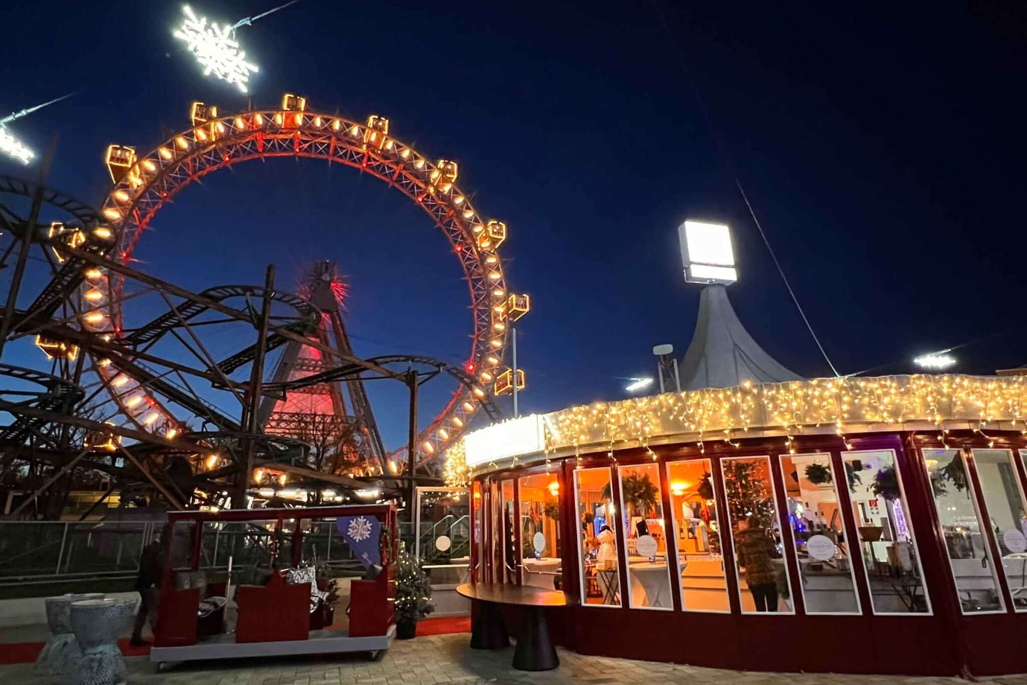 Ein hell erleuchtetes Riesenrad und eine Achterbahnanlage bei Nacht neben einem runden Gebäude, das mit festlichen Lichtern geschmückt ist.